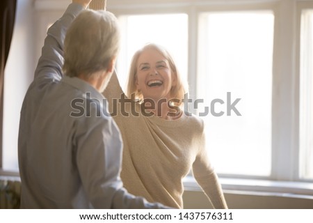 Happy middle aged mature woman enjoying dancing with elder husband at home, active healthy senior old couple man and woman pensioners having fun in waltz laughing bonding celebrating anniversary