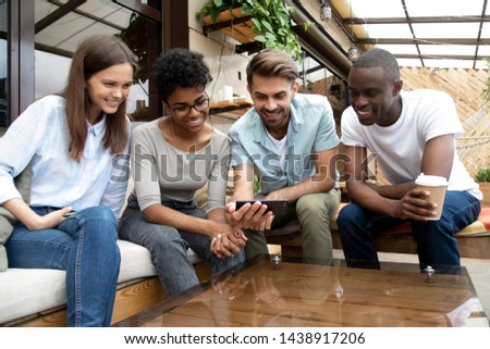 Photo of Happy multiethnic millennial friends relax in café together watch funny video on smartphone laughing, smiling diverse young people have fun in coffee shop hanging out outside. Technology concept
