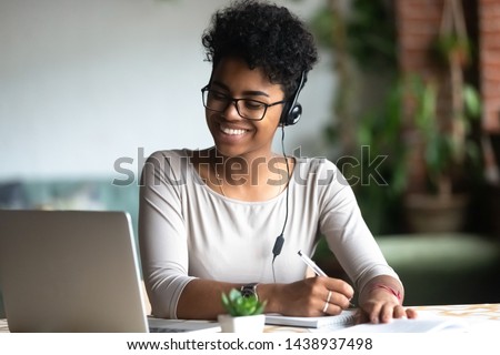 Photo of Smiling african American millennial female student in headphones and glasses sit at desk watch webinar making notes, happy biracial young woman in earphones work study using computer write in notebook