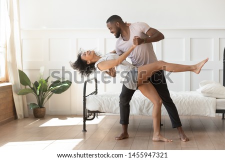 Cheerful active romantic african american couple wearing pajamas dancing in bedroom together, happy carefree young black husband and wife enjoy weekend morning laughing bonding having fun at home