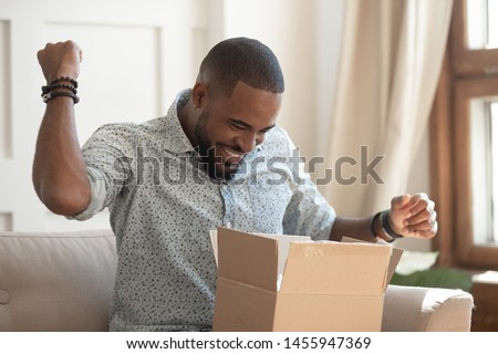 Excited african man customer receive good parcel open cardboard box at home satisfied with great purchase, happy black male consumer unpack package look inside overjoyed by postal shipping delivery