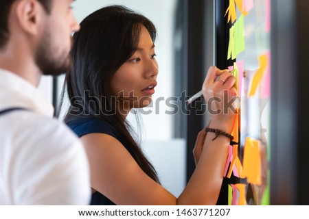 Focused asian business woman mentor coach leader writing idea or task on post it sticky notes on glass wall, serious team people developing work plan in creative corporate office at stand up meeting