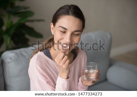 Young happy smiling mixed race woman sitting on a sofa at living room, holding glass of pure water, taking daily vitamin d, c complex, supplements for hair, skin and nail strengthen, healthcare Foto stock © 