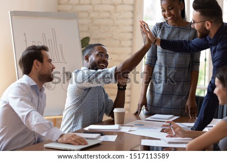 Happy african american male manager sitting at table on business team brainstorming meeting, giving high five to arabian colleague, congratulating with shared teamwork success or good project idea.