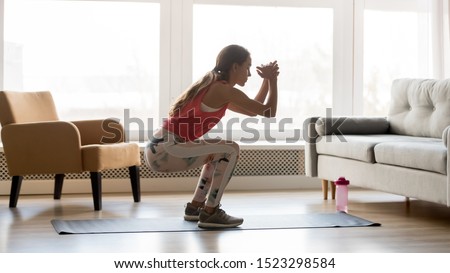 Sporty young woman doing squat morning exercise alone in living room, serious fit girl wearing sportswear crouching training muscles workout at home for healthy body lifestyle concept, side view Stock foto © 