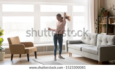 Happy carefree young woman dancing alone having fun at home listening to good music, energetic girl moving jumping in modern living room interior with large window enjoy freedom and active lifestyle