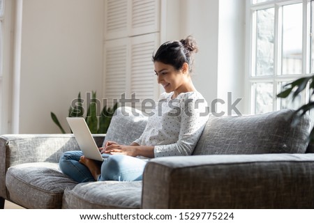 Satisfied Indian woman relaxing on comfortable couch, using laptop at home, happy girl chatting with friends in social network, spending lazy weekend, watching movie, shopping online, writing email