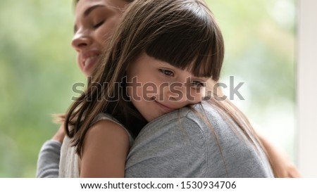 Side view close up head shot happy little adopted kid girl put head on mothers shoulder, feeling love and support. Small cute daughter hugging embracing cuddling young smiling mother at new home.