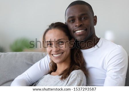 Portrait of happy international young couple hug look at camera relaxing on couch at home, loving smiling multiethnic husband and wife pose for picture together, interracial relationships concept