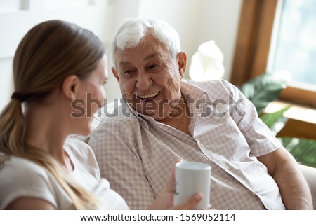 Head shot focus on happy old man talking with smiling grown up adult daughter, sitting together on sofa at home. Pleasant young woman communicating, having fun, laughing with retired 80s father.