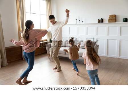 Funny active family of four young adult parents and cute small children daughters dancing together in living room interior, carefree little kids with mum dad having fun laughing enjoy leisure at home