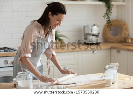 Happy attractive young adult woman lady housewife baker wear apron holding pin rolling dough on kitchen table baking pastry concept cooking cake biscuit doing bakery making homemade pizza at home
