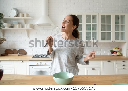 Happy young woman holding beater microphone singing song dancing cooking alone in modern kitchen, funny lady housewife having fun listening music prepare healthy morning meal doing housework at home