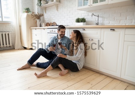 Romantic smiling couple young guy and girl sit on warm kitchen floor talking holding glasses having fun together, happy friendly husband and wife laughing enjoying drinking red wine at modern home