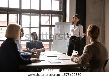 Smiling young female speaker standing near flipchart with graphs diagrams, presenting market research results to focused diverse colleagues at brainstorming meeting or educational seminar in office.