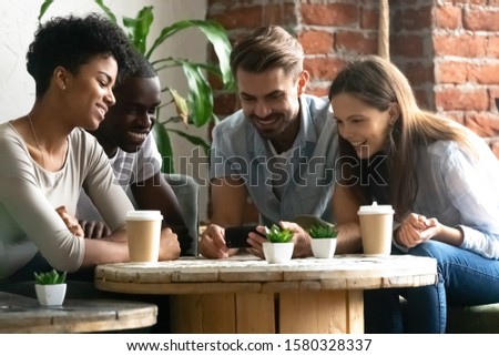 Photo of Overjoyed young multiracial people sit at table in coffeeshop smile watch funny video online on smartphone together, happy diverse millennial friends hang out in café share show pictures on cellphone