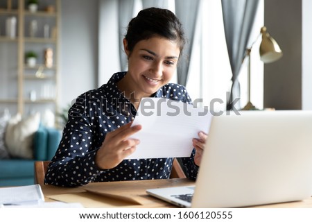 Smiling young indian woman holding paper bill letter doing paperwork bill reading good news check post mail sit at home table, happy lady customer receive bank receipt sheet tax refund notification