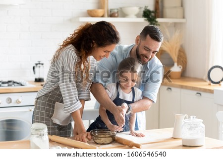 Small adorable kid girl preparing pie with loving caring parents on domestic kitchen, young family enjoy cooking process at home, teaching child, spend weekend together, help, happy parenthood concept
