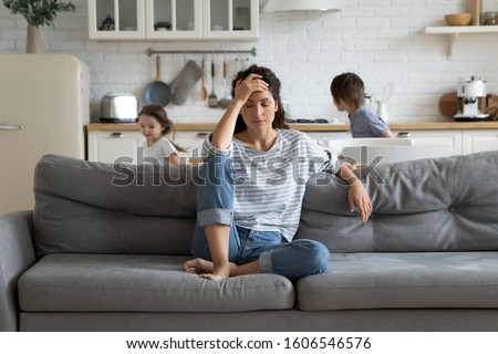 Young tired single mother suffers from headache closed eyes touch forehead sitting on couch while her daughter and son running around her and shouting, female babysitter feels exhausted by noisy kids