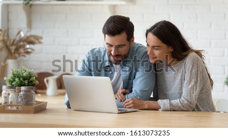 Pleasant family couple sitting at big wooden table in modern kitchen, looking at laptop screen. Happy young mixed race married spouse web surfing, making purchases online or booking flight tickets.