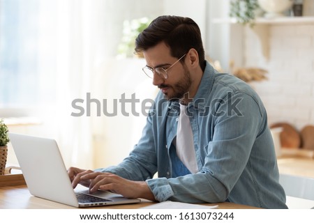 Photo of Side view handsome young businessman in eyewear working with computer remotely, sitting at wooden table in kitchen. Pleasant happy man communicating in social network, searching information online.
