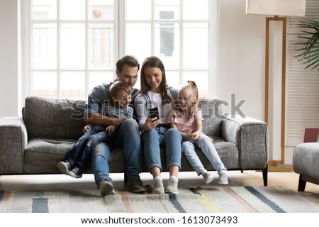 Happy young parents sit on couch with little children have fun using smartphone together, smiling caucasian family with small kids relax on sofa in living room watch funny videos on cellphone