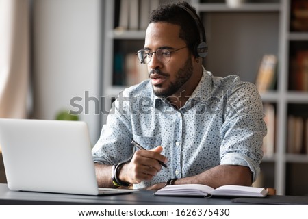 Focused young african businessman wear headphones study online watching webinar podcast on laptop listening learning education course conference calling make notes sit at work desk, elearning concept Photo stock © 