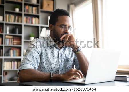 Focused concentrated young african businessman sit at desk look at laptop, serious afro american male professional analyst working online on computer data watch webinar thinking of problem solution