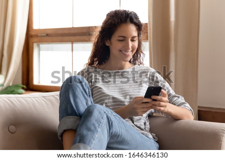 Happy pleasant millennial woman relaxing on comfortable couch, holding smartphone in hands. Smiling young lady chatting in social networks, watching funny videos, using mobile applications at home.