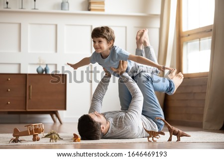 Photo of Joyful young man father lying on carpet floor, lifting excited happy little child son at home. Full length carefree two generations family having fun, practicing acroyoga in pair in living room.