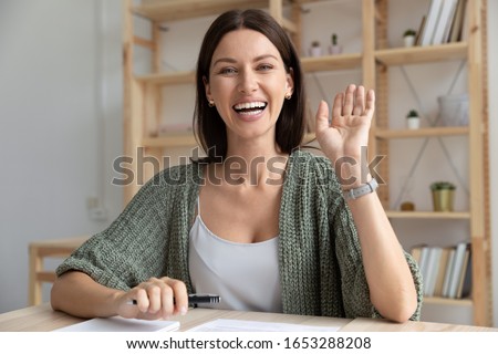 Head shot happy young 30s woman sitting at wooden desk, looking at camera, waving hello. Excited businesswoman teacher lecturer recording educational video, greeting students at online workshop.