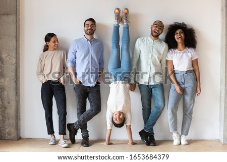 Indian girl fools around standing on hands upside-down having fun during photo shooting with multi-ethnic friends. Full length five cheery buddies laughing photographing posing near grey wall indoors 商業照片 © 