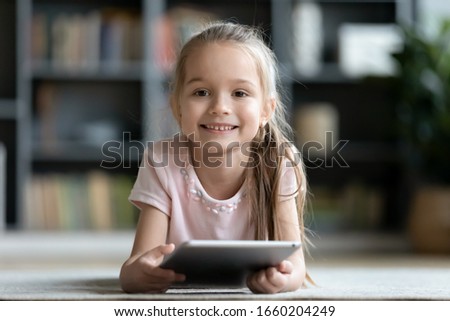 Portrait of smiling little girl lying on floor in living room using modern tablet, happy small preschooler child have fun relax at home learning or watching videos on pad device, technology concept Imagine de stoc © 