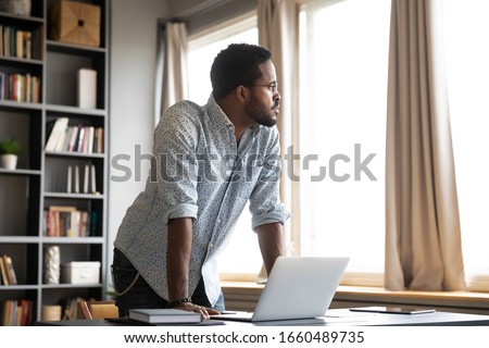 Young thoughtful african american businessman leaning on table with laptop, looking away. Millennial hipster biracial entrepreneur thinking of problem solution, future challenges in office or home.