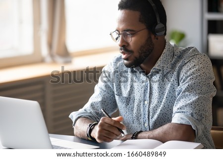 Head shot focused young african american man in glasses wearing wireless headphones, looking at laptop screen, watching educational lecture seminar webinar online, studying remotely, writing notes.