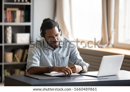 Photo of Concentrated millennial african american guy wearing earphones, listening to favorite music while planning workday. Focused young biracial businessman watching educational lecture. writing down notes.