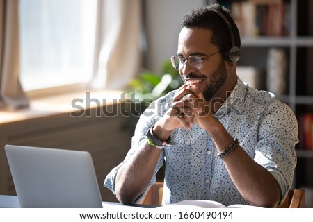 Photo of Happy millennial african american man in glasses wearing headphones, enjoying watching educational webinar on laptop. Smiling young mixed race businessman holding video call with clients partners.