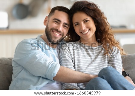 Family portrait of happy millennial husband and wife sit on couch hug cuddle look at camera posing, smiling young couple embrace show love affection, relax at home on weekend together