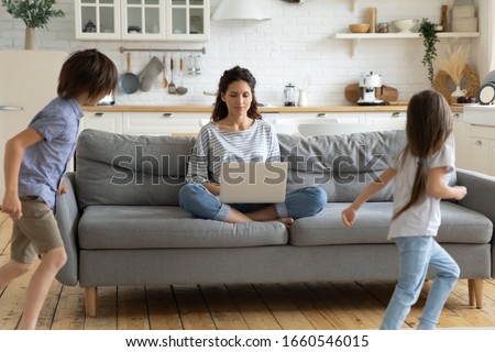 Calm young mother or nanny sit on couch working on laptop, little kids play around, peaceful mom relax on sofa use modern computer distracted from noise, parenting, upbringing concept
