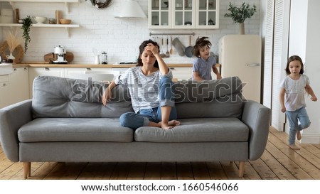 Exhausted young mum sit on couch in kitchen feel unwell tired from ill-behaved loud little children running playing, sick annoyed mother or nanny relax on sofa suffer from headache, parenting concept