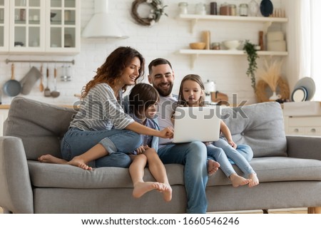 Happy young family with little kids sit on sofa in kitchen have fun using modern laptop together, smiling parents rest on couch enjoy weekend with small children laugh watch video on computer at home