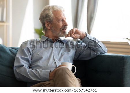 Pensive elderly 60s man sit relax on couch in living room drinking tea look in window distance thinking, thoughtful mature 50s husband rest on sofa at home feel sad pondering considering future