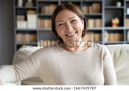 Head shot close up portrait happy healthy middle aged woman sitting on comfortable couch at home. Smiling pleasant 50s elderly mother looking at camera, posing for photo in modern living room.