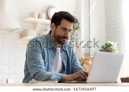 Focused young man wearing glasses using laptop, typing on keyboard, writing email or message, chatting, shopping, successful freelancer working online on computer, sitting in modern kitchen Stock foto © 