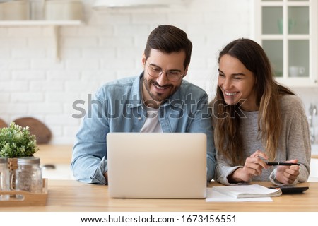 Happy young couple planning budget, reading good news in email, refund or mortgage approval, smiling woman and man looking at laptop screen, checking finances, sitting at table at home together
