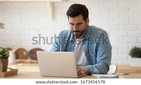 Serious man wearing glasses working on laptop online, sitting at table in kitchen, looking at computer screen, focused male using internet banking service, writing email, searching information