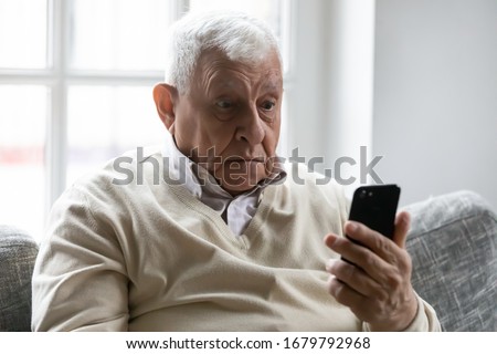 Old man sit on sofa hold smartphone look at device screen feels confused shocked by received sms message. Older generation and modern gadgets apps usage difficulties, broken cell, need repair concept Foto d'archivio © 