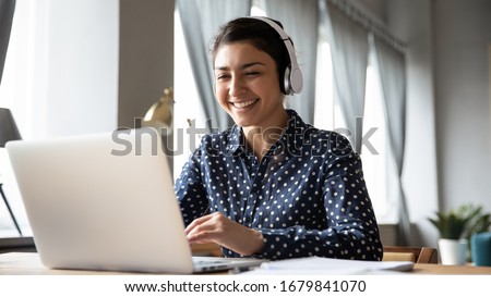 Smiling Indian girl wearing headphones using laptop, looking at screen, happy young female listening to favorite music while working online on project, excited student learning language