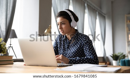 Busy young Indian woman wearing headphones working on laptop, looking at screen, making video call, engaged in conference, focused student listening to lecture, learning language online