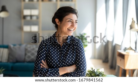 Head shot smiling Indian woman with arms crossed dreaming about good future, laughing young female standing at home, looking to aside at window, visualizing, thinking about new opportunity